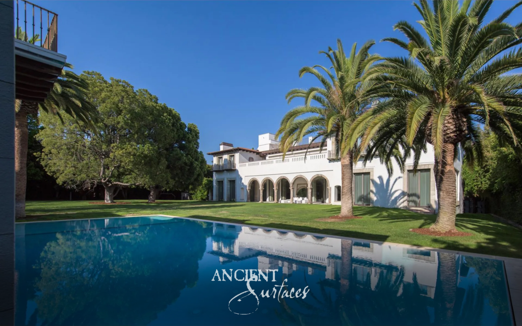 Mediterranean villa seen across the pool, its ground floor opened by an arcade of reclaimed limestone columns among palms and lawn