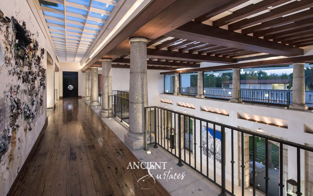 Upper gallery lined with hand hewn limestone columns carrying dark timber beams above an iron balustrade and a double height stone hall
