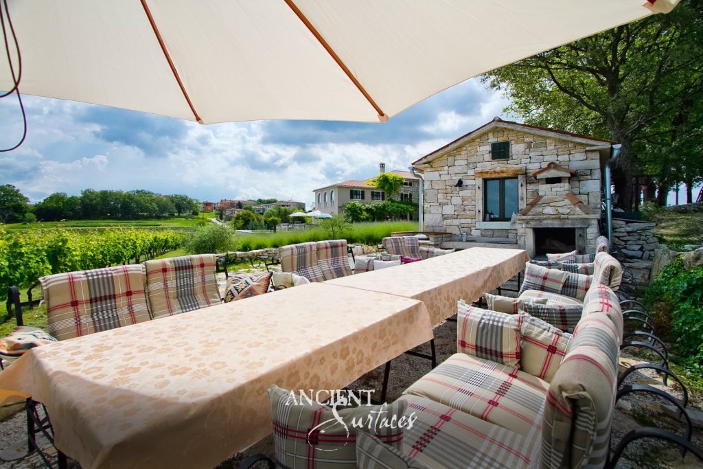 Long outdoor dining table shaded by umbrellas facing a stone cottage built by hand from irregular reclaimed limestone blocks around an outdoor oven and hearth