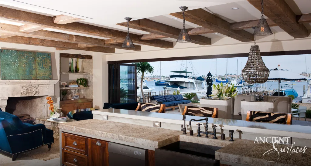 Carved limestone fireplace anchoring the open living room beyond a kitchen island topped in thick reclaimed limestone, under reclaimed beams facing the marina