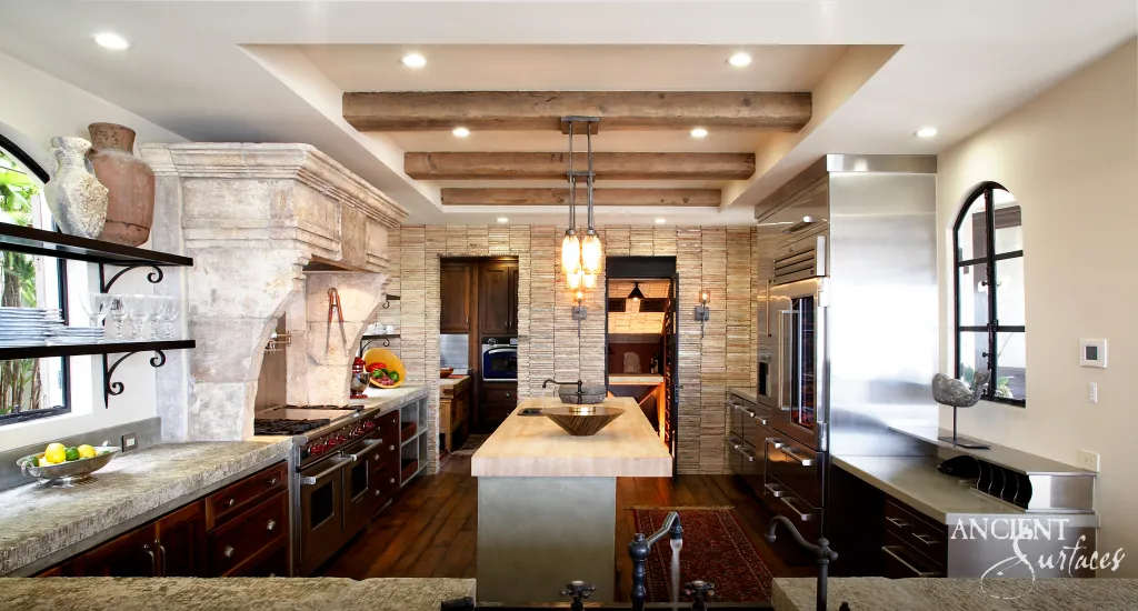 Kitchen centered on a carved reclaimed limestone hood, with thinly sliced limestone wall cladding, chiseled limestone countertops and reclaimed timber beams
