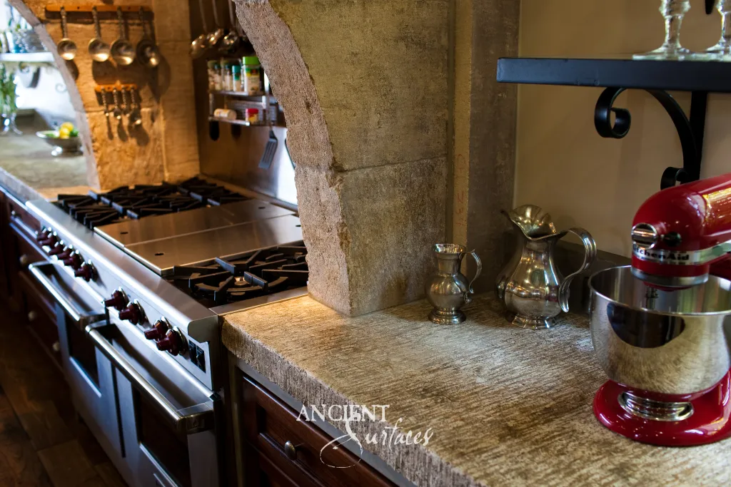 Limestone countertop chiseled by hand to a raw cut edge, its fossil flecked surface meeting the carved stone leg of the kitchen hood